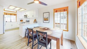 A modern kitchen with white cabinets, a central island, and a dining area with a wooden table and chairs. - Spacious home with three decks, views, game room, fireplace, & fast WiFi (Lake Arrowhead)