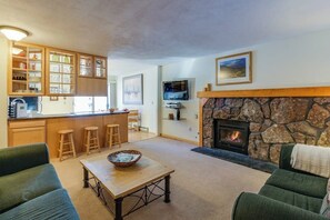 Ein Wohnzimmer mit Kamin aus Stein, eine Küche mit Holzschränken, ein Essbereich mit Barhocker und ein an der Wand befestigter Fernseher.