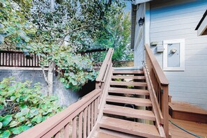 Eine Holztreppe führt zu einem Haus mit hellblauer Fassade, einer weißen Tür und einem kleinen Garten mit viel Grün.