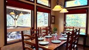 Comedor con mesa de madera dispuesta para cuatro personas, sillas alrededor y amplias ventanas que dan a vistas de árboles y una terraza exterior.
