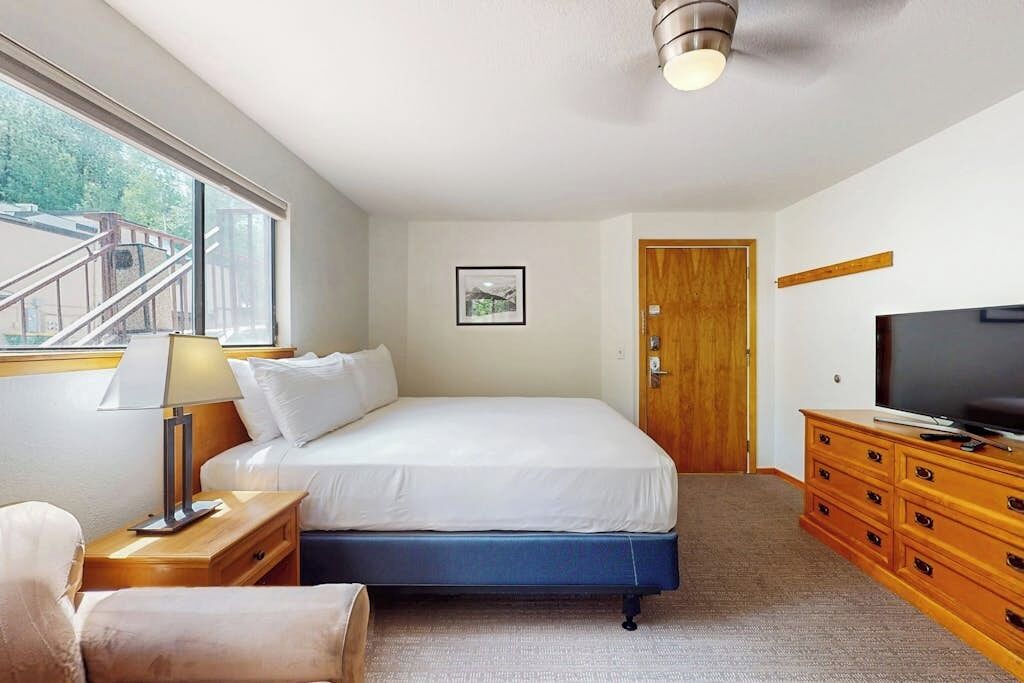 Un dormitorio con una cama, televisión, cómoda de madera, mesita de noche, lámpara, un cuadro enmarcado y una puerta.
