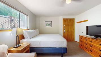 Un dormitorio con una cama, televisión, cómoda de madera, mesita de noche, lámpara, un cuadro enmarcado y una puerta.