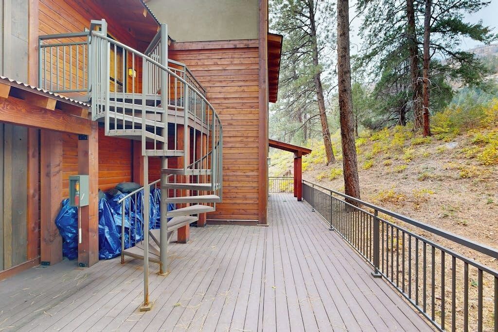A wooden cabin with a wooden deck, a metal staircase, and a view of a forested area.