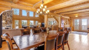 Un intérieur spacieux de chalet en bois, avec une grande table à manger, des chaises en bois, un lustre, une cheminée en pierre et un escalier menant à un balcon.