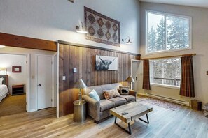 Ein gemütliches Wohnzimmer mit Holzboden, einer Couch, einem Couchtisch und einem großen Fenster mit Vorhängen.