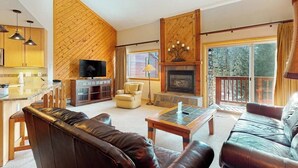 Ein Wohnzimmer mit Kamin, Ledersofas, einem Fernseher und einer Küchenzeile mit Holzwänden und Pendelleuchten.