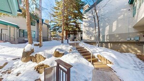A snow-covered outdoor area with steps leading to a building entrance, surrounded by trees and rocks. - Cozy condo w/ shared pool, hot tub & full kitchen - walk to lifts (Snowmass Village)