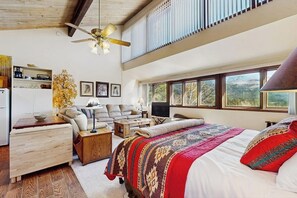 A spacious bedroom with a large bed, a ceiling fan, a television, and a view of the outdoors through large windows. - Studio condo w/ wifi, kitchenette, shared pool & hot tub - near golf & skiing (Durango)