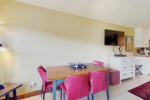 A dining area with a wooden table and pink chairs, a white cabinet with a flat-screen TV, and a kitchen area with white cabinets and a red rug. - Spacious studio with lovely view and access to shared pool and hot tub (Durango)
