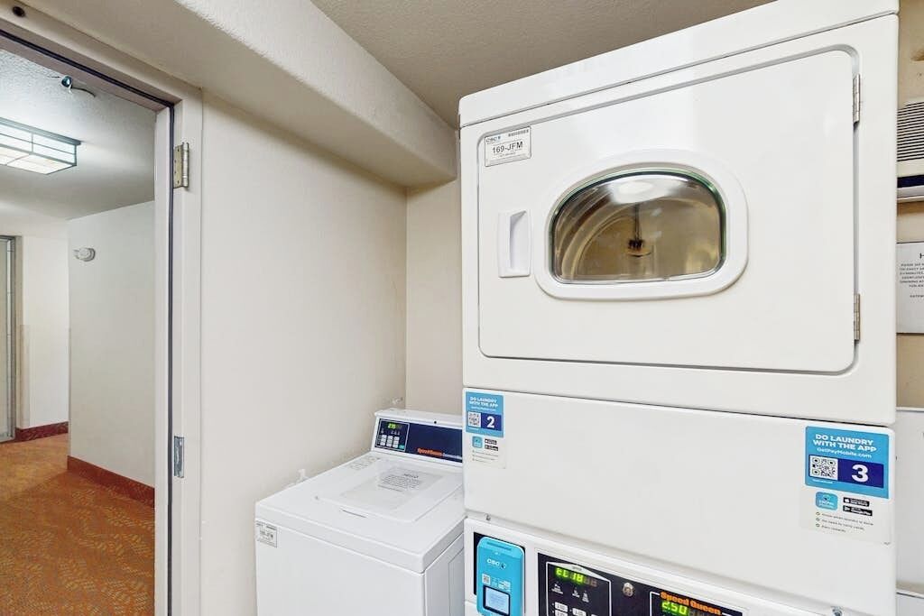 Un espace buanderie équipé d’une pile de lave-linge et de sèche-linge, d’un panneau de commande numérique et d’une porte donnant sur une autre pièce.