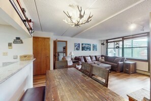 Ein modernes Wohnzimmer mit einem Holzesstisch, einer Sofagarnitur und einem großen Fenster mit Tageslicht.