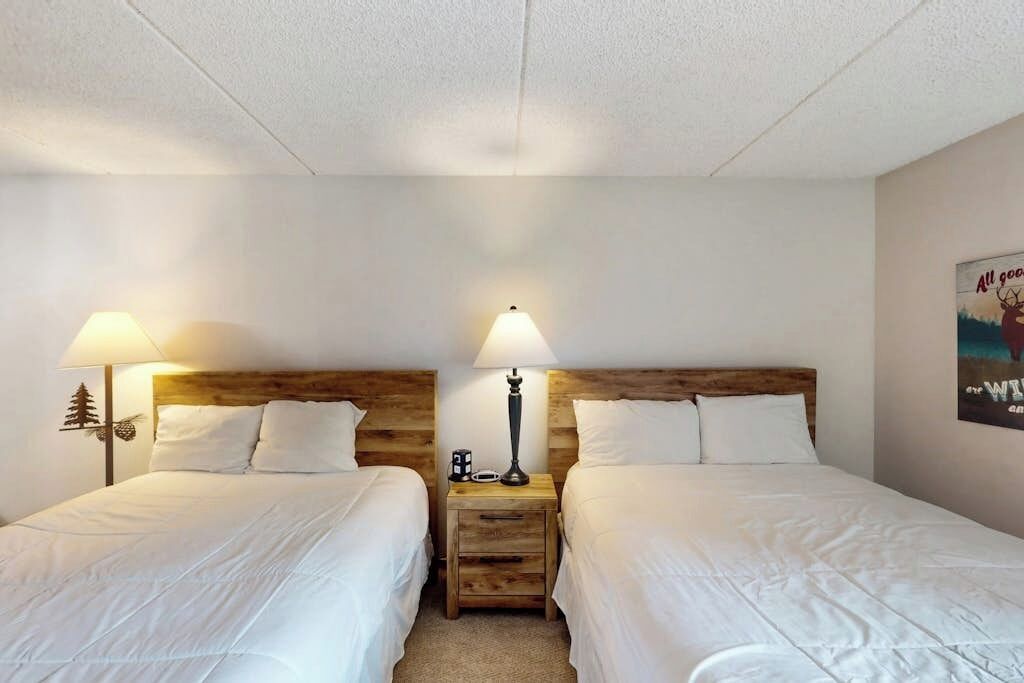 A hotel room with two beds, wooden headboards, bedside lamps, a nightstand, and a poster on the wall.