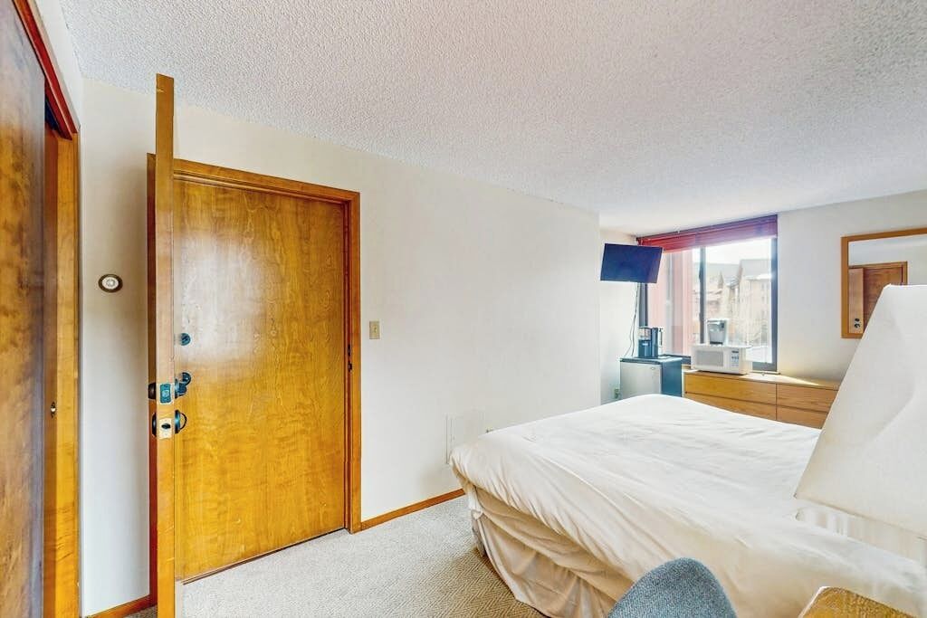 A bedroom with a wooden door, a bed with white bedding, a bedside table, a lamp, and a window with a view.