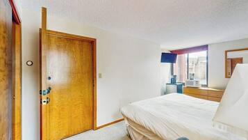 A bedroom with a wooden door, a bed with white bedding, a bedside table, a lamp, and a window with a view.
