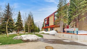 Um edifício moderno com fachada vermelha e bege, uma placa com a inscrição 'VILLAGE SQUARE' e uma área pavimentada com montes de neve e árvores.