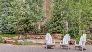Three Adirondack chairs, a stone wall, and a variety of trees in a landscaped outdoor area. - Family-friendly mountain condo w/balcony, shared hot tub, & mountain views (Copper Mountain)