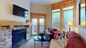 A living room with a stone fireplace, a television mounted on the wall, a red sofa, and large windows with blinds. - Family-friendly mountain condo w/balcony, shared hot tub, & mountain views (Copper Mountain)