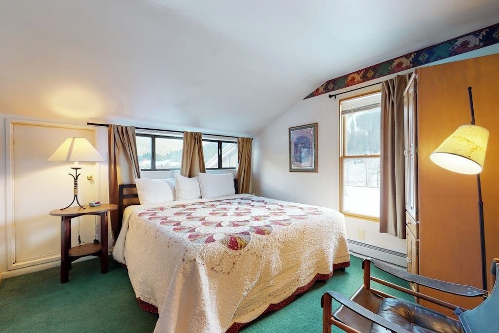 Une chambre d’hôtel avec un lit, une chaise, une lampe, une fenêtre avec des rideaux et un tableau au mur.
