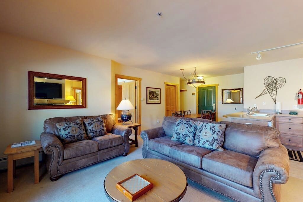A living room with two sofas, a coffee table, a TV, and a dining area in the background.