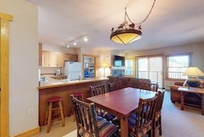A dining area with a wooden table and chairs, a kitchen with wooden cabinets, a bar area, and a living room with a sofa and a lamp. - Chalet next to gondola w/shared fitness center, hot tub, & sauna (Copper Mountain)