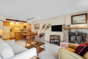 A living room with a sofa, coffee table, fireplace, television, and a kitchen area with wooden cabinets and a dining set. - Mountain view condo w/gas fireplace, shared outdoor pool, and free WiFi (Copper Mountain)