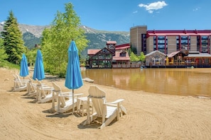 A lakeside setting with Adirondack chairs and blue umbrellas, a wooden cabin, and a modern building with a red roof. - Ski-in ski-out townhouse w/mountain views, gas fireplace, WiFi (Copper Mountain)