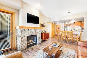 Ein Wohnzimmer mit Kamin aus Stein, einem an der Wand befestigten Fernseher, einem Essbereich mit Holzsesseln und einer Küche mit modernen Geräten.