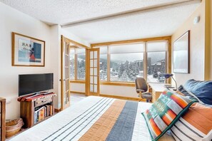 Ein Schlafzimmer mit Bett, Fernseher, Schreibtisch und Blick auf eine verschneite Landschaft durch große Fenster.