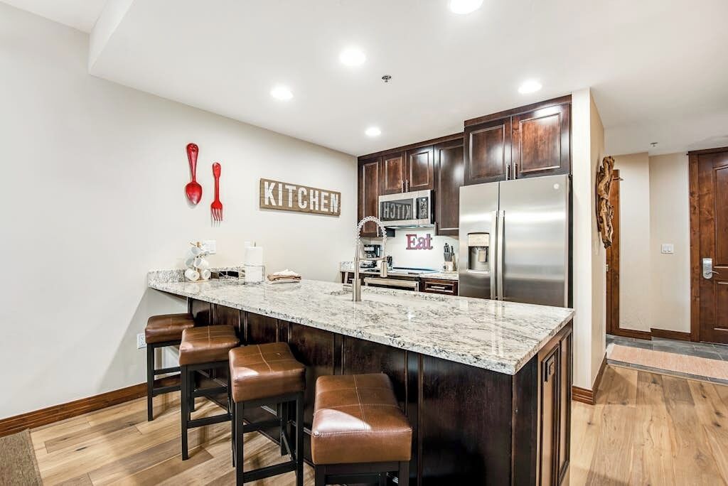 A modern kitchen with a marble countertop, dark wooden cabinets, a stainless steel refrigerator, and a 'KITCHEN' sign on the wall.