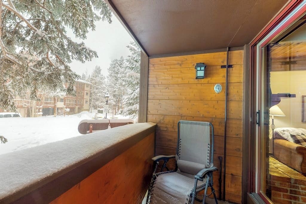 Una cabaña de madera con balcón, una silla y una ventana con vistas a árboles nevados y edificios.