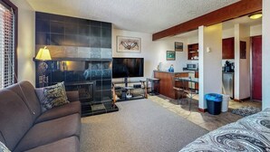 A living room with a sofa, a fireplace, a television, and a kitchen area with a microwave and cabinets. - Riverfront studio with mtn view, balcony, & fireplace (Breckenridge)