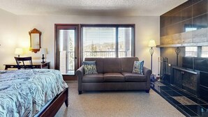 A hotel room with a bed, a sofa, a fireplace, and large windows. - Riverfront studio with mtn view, balcony, & fireplace (Breckenridge)