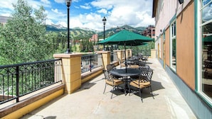 Eine Terrasse im Freien mit Tischen und Stühlen, einem grünen Sonnenschirm und Blick auf Berge.