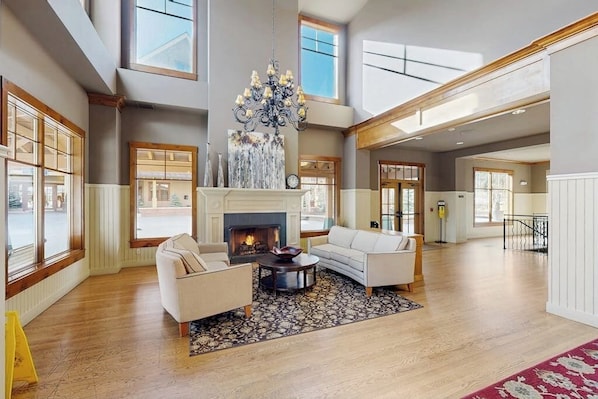 A spacious living room with a fireplace, large windows, and a high ceiling. - Modern condo, shared hot tub, steps to QuickSilver Super Chair lift (Breckenridge)