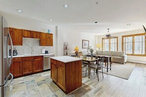 A modern kitchen with wooden cabinets, a marble countertop, and a dining area with a table and chairs. - Modern condo, shared hot tub, steps to QuickSilver Super Chair lift (Breckenridge)