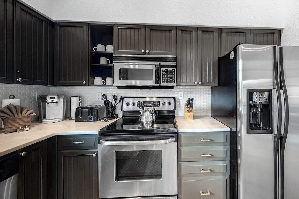 A modern kitchen with dark cabinets, stainless steel appliances, a kettle, and a coffee maker.