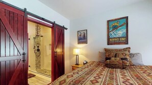 Una habitación con cama, puerta de madera, ducha, un cartel y mesita de noche con lámpara.