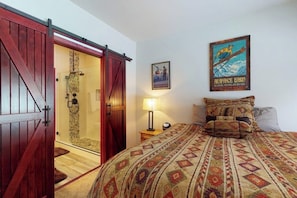 A room with a bed, a wooden door, a shower, a poster, and a nightstand with a lamp. - Modern Keystone condo w/ a gas fireplace & shared hot tub (Keystone)