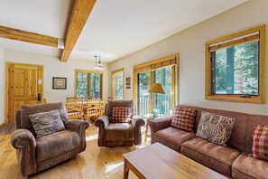 A spacious living room with a wooden ceiling, large windows, a brown leather sofa, a brown leather armchair, and a wooden coffee table. - Delightful condo with hot tub, gas fireplace & heated garage (Breckenridge)