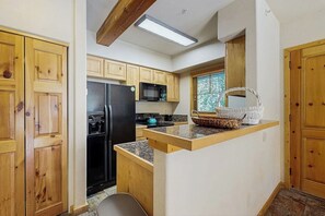 A kitchen with wooden cabinets, a black refrigerator, a microwave, and a window with a view of trees. - Delightful condo with hot tub, gas fireplace & heated garage (Breckenridge)