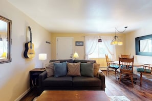 Ein Wohnzimmer mit Sofa, Couchtisch, Gitarre, Lampe sowie Essbereich mit Stühlen.