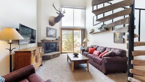 A living room with a fireplace, a mounted elk head, a staircase, and a television. - Third-Floor Condo + Loft w/Mtn Views, Balcony, Wood Fireplace, & Shared Hot Tub (Copper Mountain)
