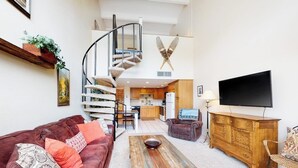 A living room with a sofa, coffee table, TV, wooden cabinet, and a spiral staircase leading to a kitchen. - Third-Floor Condo + Loft w/Mtn Views, Balcony, Wood Fireplace, & Shared Hot Tub (Copper Mountain)