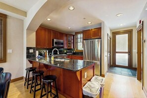 A kitchen with wooden cabinets, a stainless steel refrigerator, a microwave, and a large island with stools. - Cozy townhome with fully stocked kitchen, fast wifi & easy ski access (Copper Mountain)