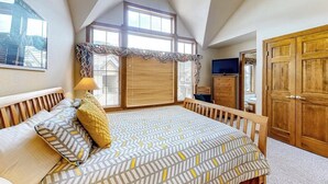 Ein Schlafzimmer mit schrägen Wänden, einem Bett mit gemusterten Textilien, einem Fenster mit Jalousien, einem Holzschrank und einem Fernseher.