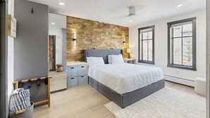 A modern bedroom with a large bed, a built-in bench, a mirror, and a stone wall. - Charming, walk-to-lifts condo with hot tubs, pool, gym & gas fireplace (Breckenridge)