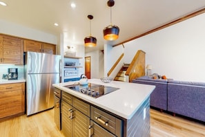 A modern kitchen with a stainless steel refrigerator, wooden cabinets, a central island, pendant lighting, and a staircase leading to a living area with a sofa.