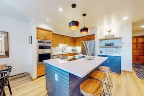 A modern kitchen with wooden cabinets, stainless steel appliances, a central island with stools, and pendant lighting.
