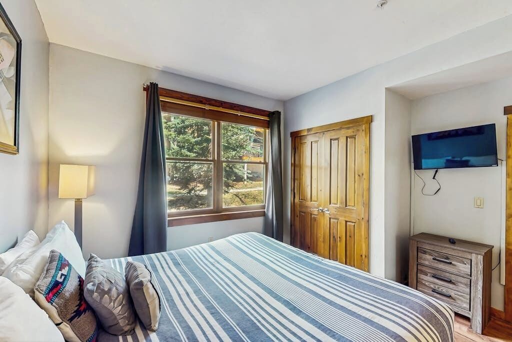 A bedroom with a bed, a TV, a window with curtains, and a wooden door.