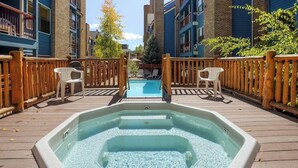 Ein Whirlpool auf einer Holzterrasse mit einem Pool im Hintergrund, umgeben von Gebäuden.